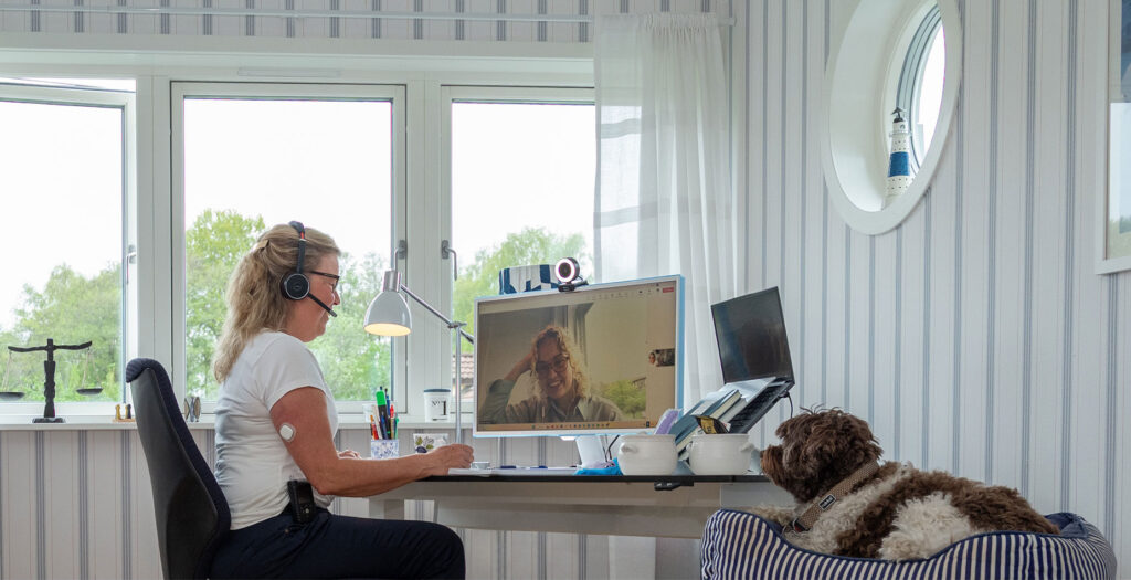 En kvinna som bär ett headset sitter vid ett skrivbord och har ett videosamtal med support i diabetes, på sin dator, medan en hund ligger i en randig säng i närheten på ett ljust hemmakontor.