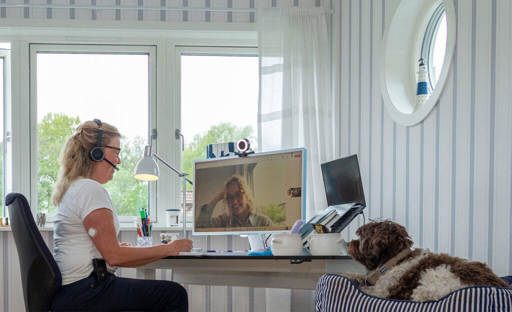 En kvinna som bär ett headset sitter vid ett skrivbord och har ett videosamtal med support i diabetes, på sin dator, medan en hund ligger i en randig säng i närheten på ett ljust hemmakontor.